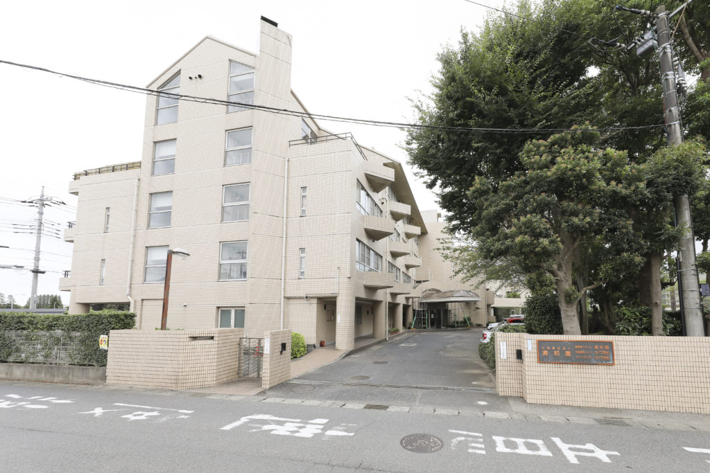 介護施設の建物外観。5階建てのベージュタイル張りの建築。エントランス前の車寄せとアプローチが見える。